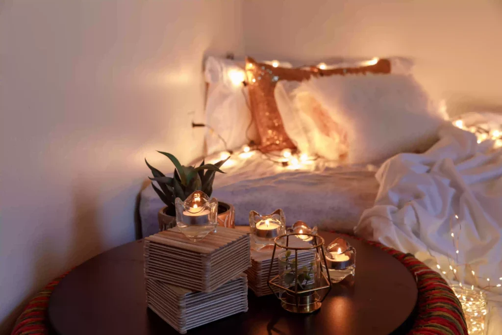 A cozy bed with a fairy lights and bedside table decorated by candle lights and small plants.
