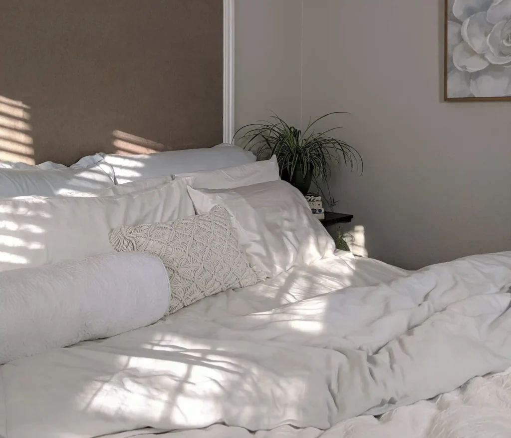 A cozy bed with white bedsheet and pillow beside is table while hitting by a sun ray.